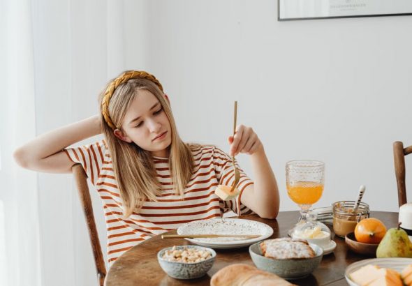 Teen girl in striped shirt at breakfast table with fruits, bread, and juice. Casual morning setting.