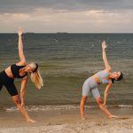 Two women exercising at a beach, performing yoga poses with the ocean in the background during sunset.