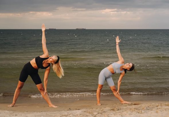Two women exercising at a beach, performing yoga poses with the ocean in the background during sunset.